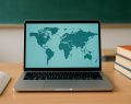 A laptop displaying a map of the world on a wooden desk with stacked books and an open notebook nearby.