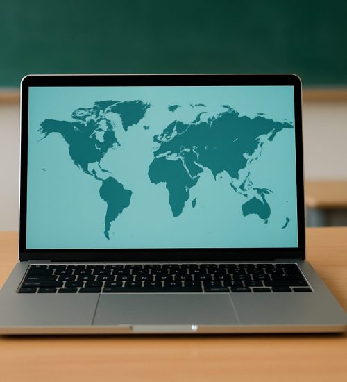 A laptop displaying a map of the world on a wooden desk with stacked books and an open notebook nearby.